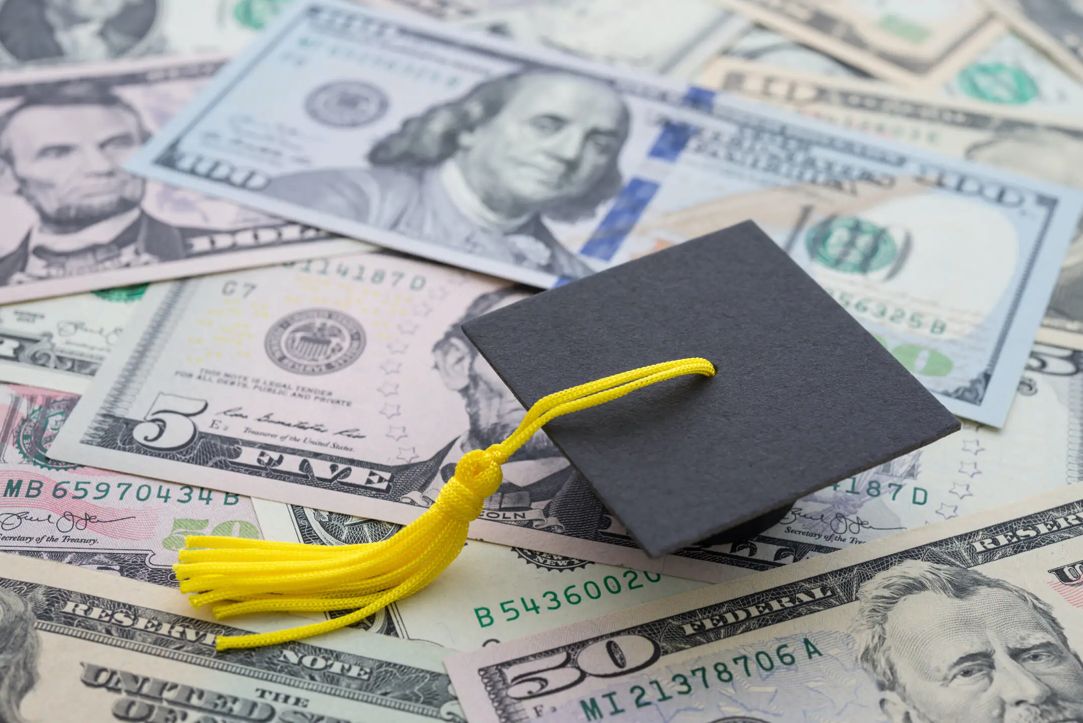 A paper graduation cap is sitting on a bunch of paper money laid out flat showcasing types of student loans available.