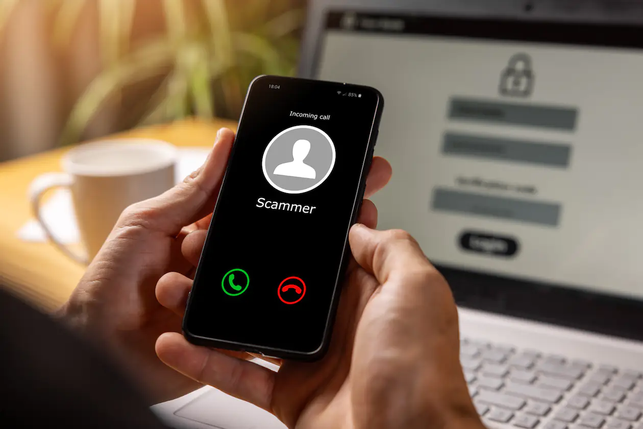 A person is holding a cell phone with an incoming call and the word scammer on the screen indicating bank fraud.