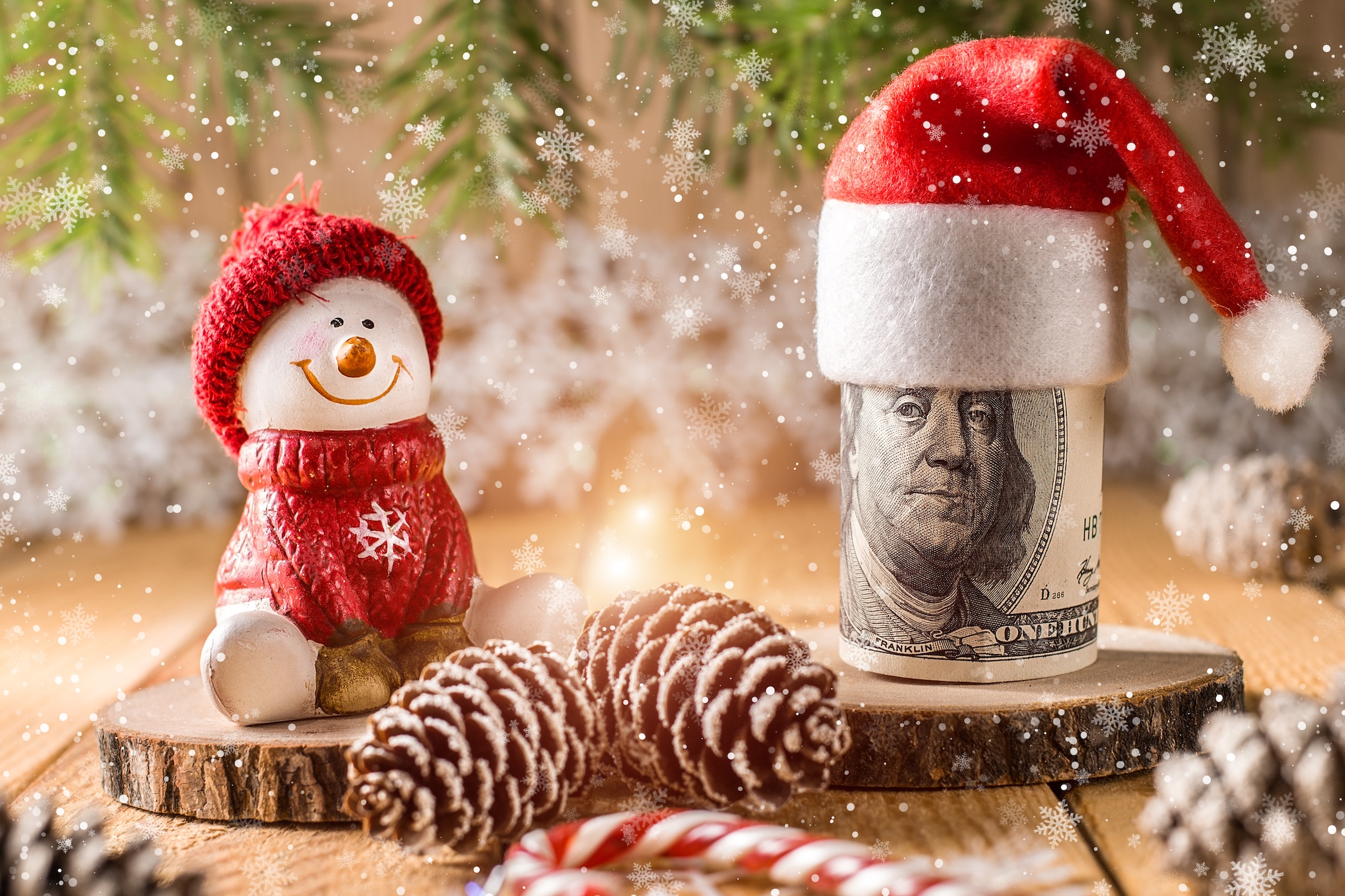 A snowman figurine is sitting by rolled up cash with a santa hat on it showcasing holiday budgeting.