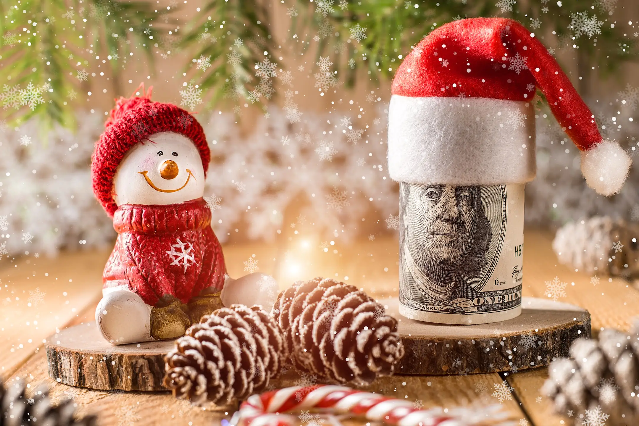 A snowman figurine is sitting by rolled up cash with a santa hat on it showcasing holiday budgeting.